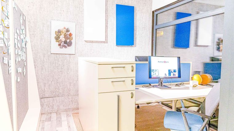 Ein modernes Büro mit einem Schreibtisch, zwei Bildschirmen und einem Rollcontainer. Die Wände sind mit blauen Akzenten und Kunstwerken dekoriert.