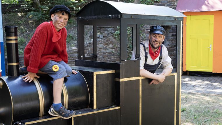 Ein Junge und ein Mann sitzen in einer Spielzeuglokomotive. Beide haben lächelnde Gesichter und tragen bunte Kleidung.