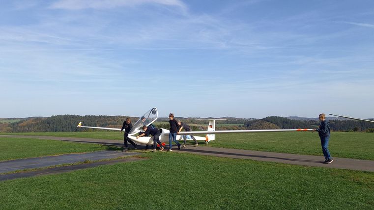 Eine Gruppe von Personen arbeitet an einem Segelflugzeug auf einem grünen Feld. Der Himmel ist klar und blau.