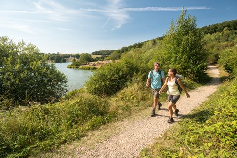 Twee wandelaars op het pad naast een meer rechts en links van het pad groene struiken, heldere lucht