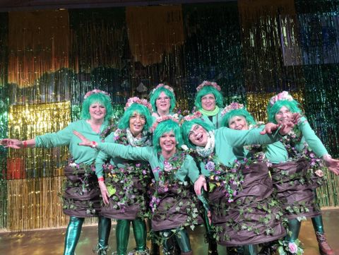 Een groep vrouwen in groene kostuums die eruitzien als planten, poseert vrolijk op het podium. Ze hebben bloemen in hun haar en lachen voor de foto.