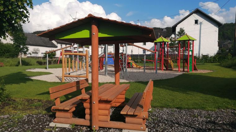 Ein Spielplatz mit bunten Spielgeräten und einer überdachten Sitzgelegenheit. Im Hintergrund sind grüne Wiesen und Häuser zu sehen.