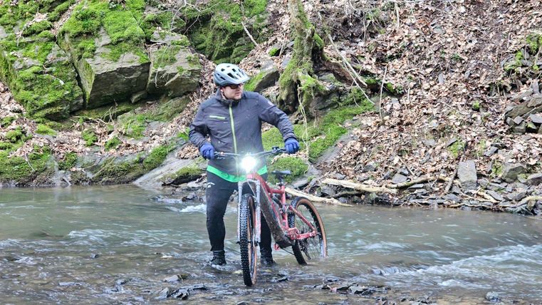 A cyclist crosses a narrow, flowing stream in a wooded area. In the background, moss-covered rocks and dry leaves are visible.