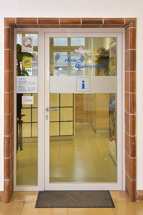 Ein Empfangsbereich mit gläserner Tür und Informationsschild. Die offene Tür lässt einen einladenden Raum hinter der Tür erahnen.