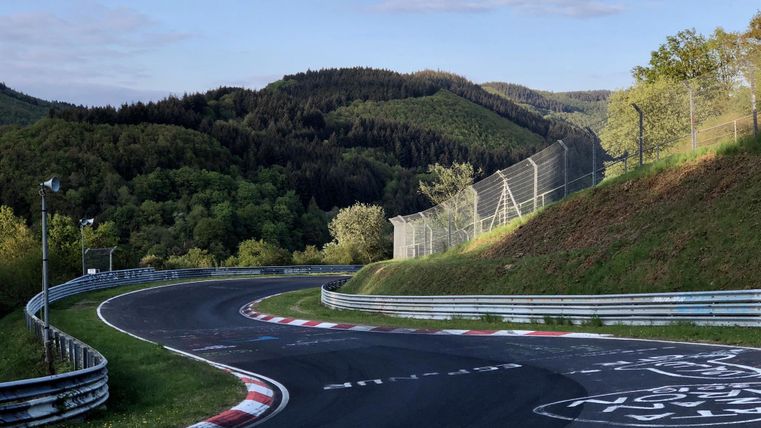 Eine kurvenreiche Rennstrecke in einer grünen Hügellandschaft. Im Hintergrund sind Bäume und ein klarer Himmel zu sehen.