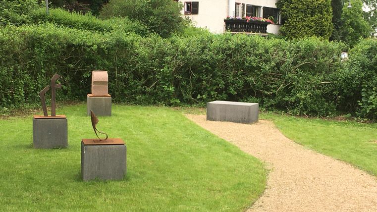Un jardin bien entretenu avec des sculptures sur des socles en pierre. Un chemin en gravier traverse l'herbe verte et la haie environnante.