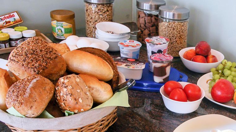 Ein reichhaltiges Frühstücksbuffet mit Brötchen, Joghurt, frischem Obst und verschiedenen Marmeladen. Die Auswahl umfasst auch Eier und Cerealien.