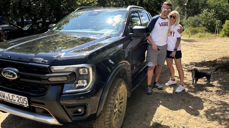 Ein Paar steht neben einem schwarzen Ford SUV auf einem unbefestigten Parkplatz. Im Hintergrund sind Bäume zu sehen.