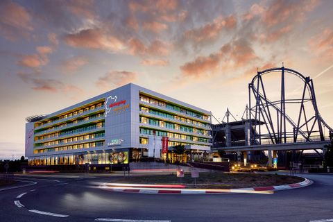A modern hotel building with colorful windows at a roundabout. In the background, a roller coaster can be seen, while the sky glows in evening colors.