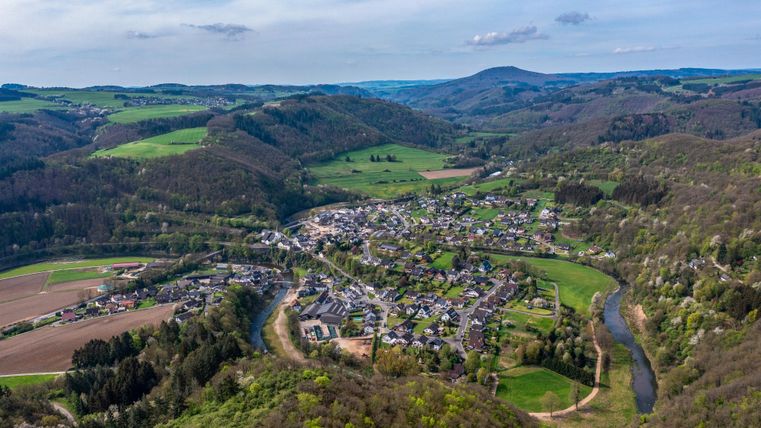 Eine malerische Landschaft mit einem kleinen Dorf, umgeben von sanften Hügeln und Wiesen. Ein Fluss schlängelt sich durch die grüne Umgebung.