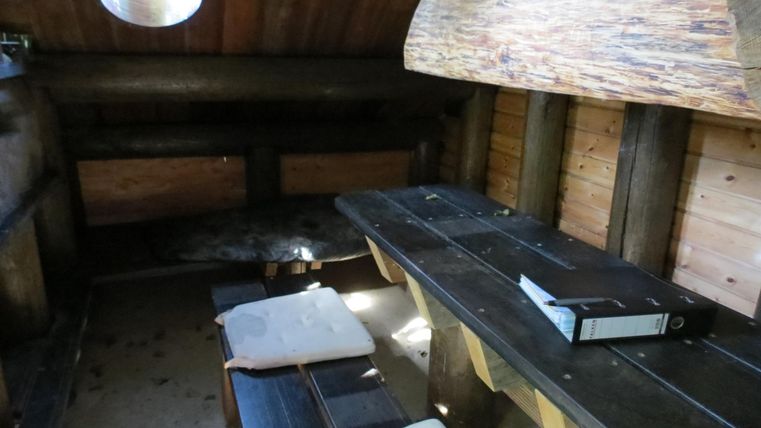 Ein rustikaler Raum mit Holzwänden und einem langen Tisch. Auf der linken Seite sind Bänke und kleine Kissen zu sehen.