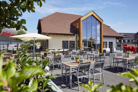 Un restaurant moderne avec de grandes fenêtres et un espace extérieur accueillant. Des tables avec des chaises sont prêtes sous des parasols.