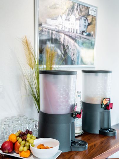 Twee sapdispensers staan op een houten tafel, daarnaast een kom met vers fruit. Op de achtergrond hangt een afbeelding van een schilderachtig landschap.