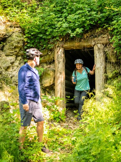 Zwei Personen stehen vor einer Höhle in der Natur. Die Umgebung ist grün mit buntem Pflanzenbewuchs, und eine Person trägt einen Fahrradhelm.