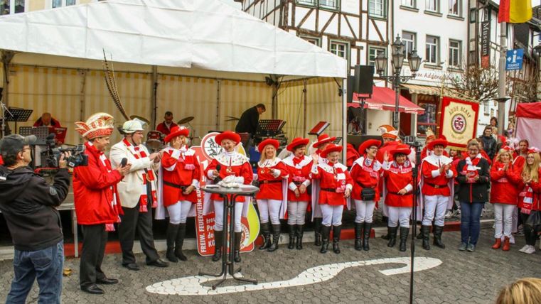 Eine Gruppe von Menschen in roten Uniformen steht auf einer Bühne und singt. Im Hintergrund sind historische Gebäude und ein Zeltdach zu sehen.