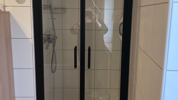 Een moderne douche met glazen front en zwarte frames. De wanden zijn betegeld en er ligt een antislip badmat op de vloer.