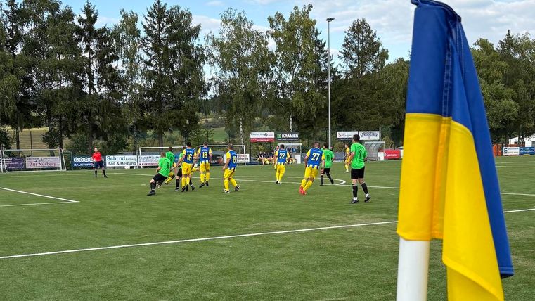 Ein Fußballspiel auf einem grünen Platz mit Spielern in gelb-blauen Trikots. Im Vordergrund weht eine Flagge in den Farben Blau und Gelb.
