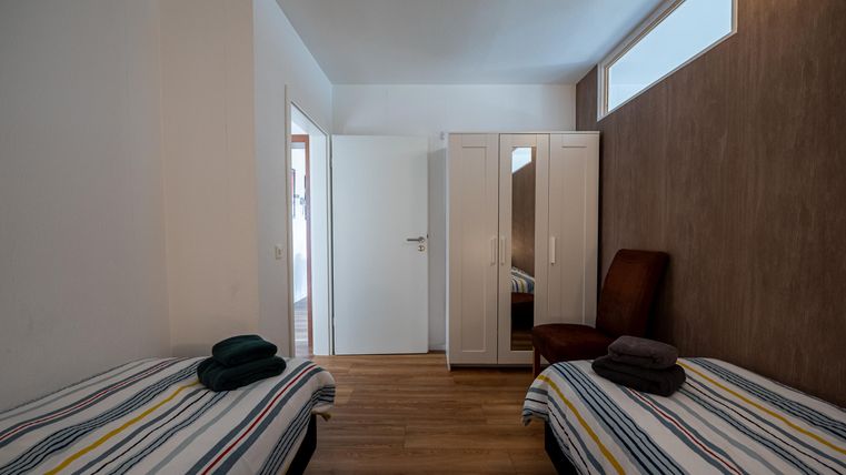 Een eenvoudige kamer met twee eenpersoonsbedden en een kast. De muren zijn licht, en het licht valt door een raam.