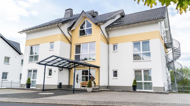 Een modern huis met een gele gevel en grote ramen. De voordeuroverkapping en de gebogen trap zorgen voor een uitnodigende indruk.