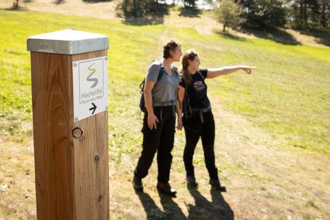 Zwei Personen auf einem Wanderweg in der Hocheifel, eine zeigt in die Ferne. Ein Wegweiser mit der Aufschrift 'Hocheifel Panorama Weg'.