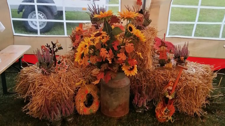 Eine herbstliche Dekoration mit Heuballen und bunten Blumen, darunter Sonnenblumen. Ein rustikales Gefäß ergänzt die Stimmung.