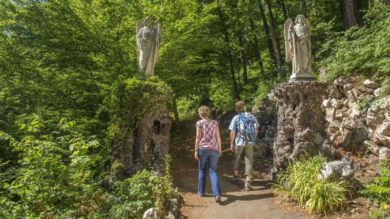 A beautiful forest path with two angel statues on the side. The visitors walk relaxed through the green surroundings.
