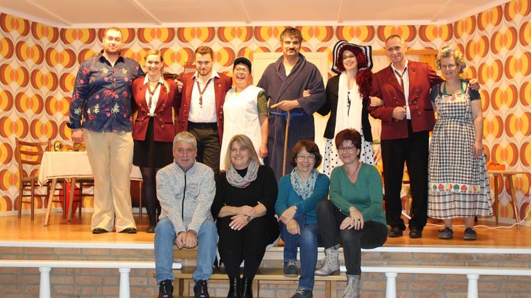 A group of people stands and sits on a stage. They are wearing different costumes and smiling at the camera.