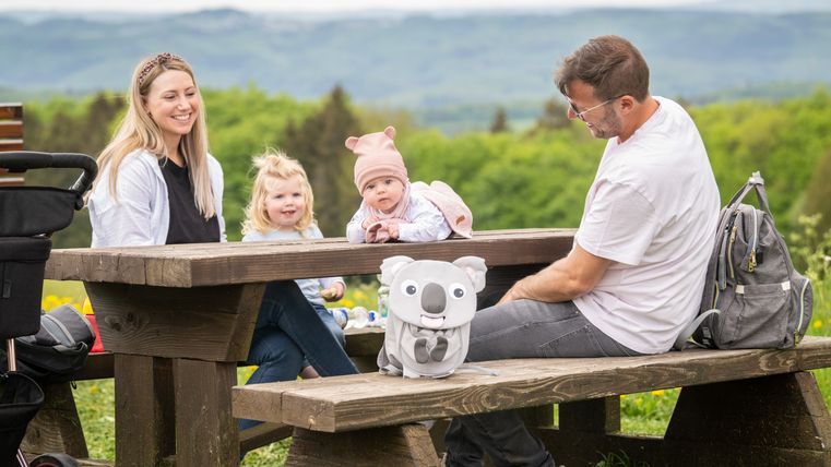 Familie bei einem Picknick auf einer Holzbank in der Natur.