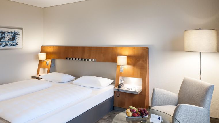 A modern hotel room with two single beds and tasteful decor. Next to the beds is a cozy armchair and a glass table with fresh fruit.