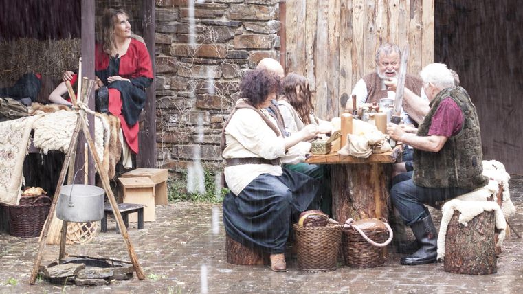 Eine Gruppe von Menschen sitzt an einem Tisch im Freien und arbeitet an handwerklichen Tätigkeiten. Im Hintergrund steht eine hölzerne Wand und es regnet leicht.