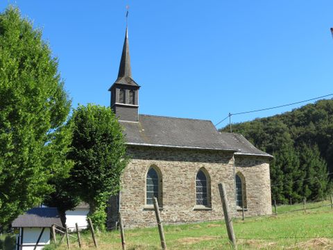 Een kleine stenen kerk met een spitze toren omgeven door bomen. De lucht is helder en blauw.