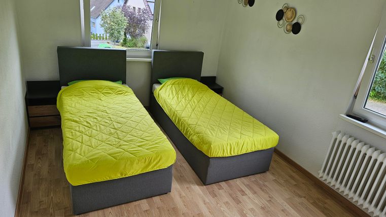 A simple bedroom with two single beds in gray and bright yellow bed covers. The room has a light wood floor and a window with a view to the outside.