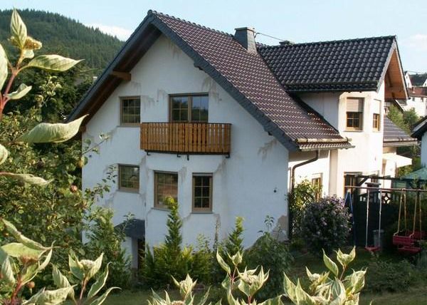 A modern house with a balcony, surrounded by green plants. In the background, gentle hills can be seen.