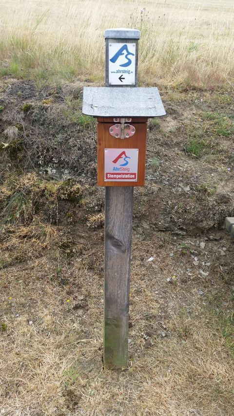 Ein Wegweiser auf einem Holzpfosten mit einem blauen und einem orangefarbenen Schild. Im Hintergrund liegt eine trockene Wiese.