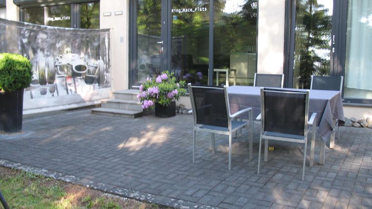 Ein schöner Außenbereich mit einem Tisch und Stühlen auf einer Terrasse. Die Gegend ist mit Pflanzen geschmückt und hat große Fenster im Hintergrund.