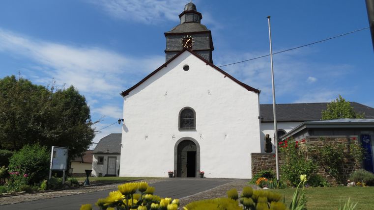 Eine weiße Kirche mit einem charakteristischen Dach und einer Uhr. Blumenbeete umgeben das Gebäude und der Himmel ist klar blau.