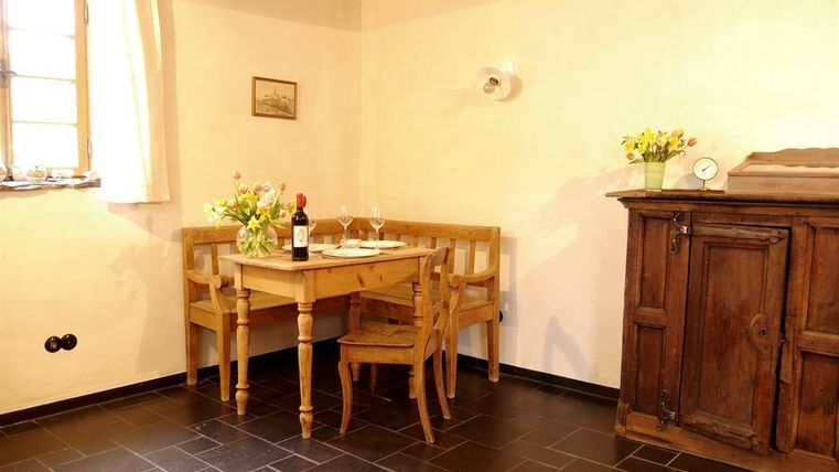 A cozy dining area with a wooden chair and table, decorated with fresh flowers. In the background stands an antique cupboard and a wine bottle is on the table.