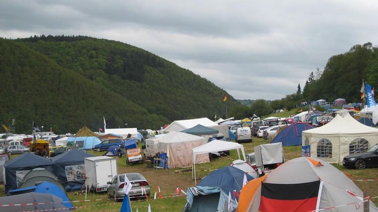 Un campement dans un paysage verdoyant avec de nombreuses tentes colorées et des véhicules. Le ciel est nuageux et il y a quelques arbres en arrière-plan.