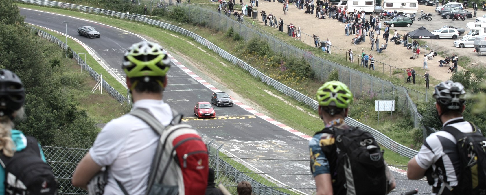 Mountain bikers watch a car race from a hill at the Br&uuml;nnchen on the N&uuml;rburgring racetrack. Spectators and motorhomes in the background., &copy; schanze communication
