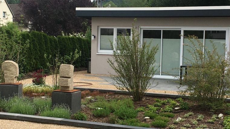 Un jardin moderne avec du gravier noir, des plantes vertes et deux sculptures en pierre. À l'arrière-plan se trouve un bâtiment avec de grandes fenêtres et un espace extérieur lumineux.