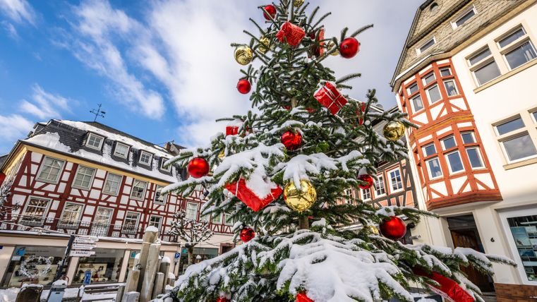 Ein geschmückter Weihnachtsbaum mit Schnee bedeckt, steht vor Fachwerkhäusern in Adenau, unter blauem Himmel.