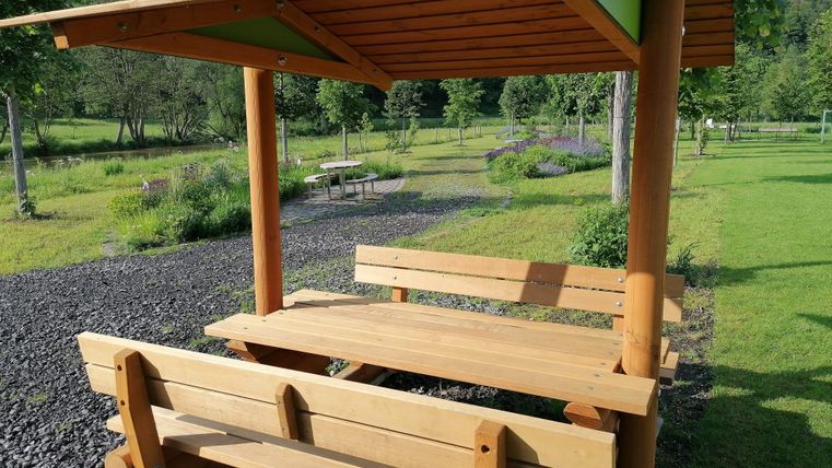 Ein schöner Holztisch mit Bänken und einem Dach steht in einer grünen Umgebung. Im Hintergrund sind Bäume und eine gut gepflegte Wiese zu sehen.