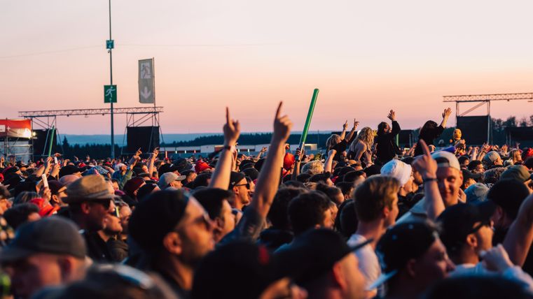 Eine große Menschenmenge bei einem Konzert, viele Hände sind in die Höhe gestreckt. Der Himmel zeigt schöne Farbtöne bei Sonnenuntergang.