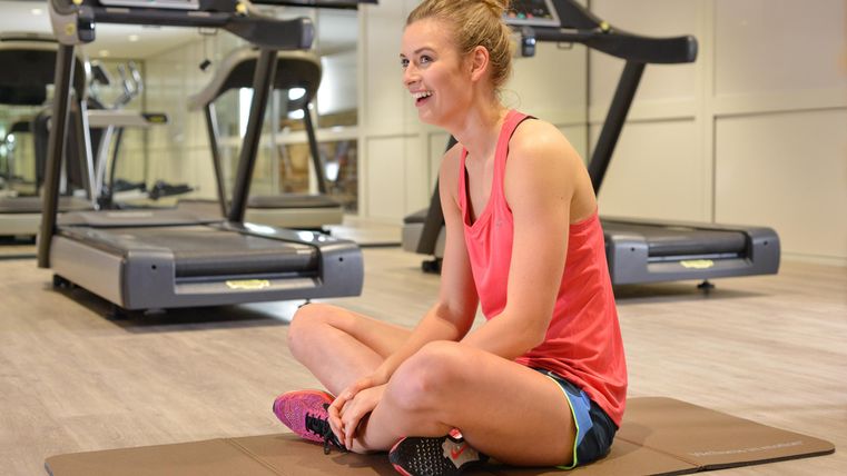 A cheerful woman is sitting on an exercise mat in a gym. Treadmills can be seen in the background.