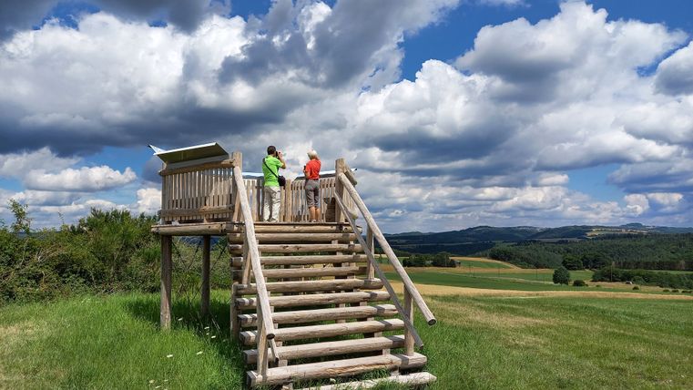 Zwei Personen stehen auf einer hölzernen Aussichtsplattform in einer ländlichen Landschaft mit bewölktem Himmel.