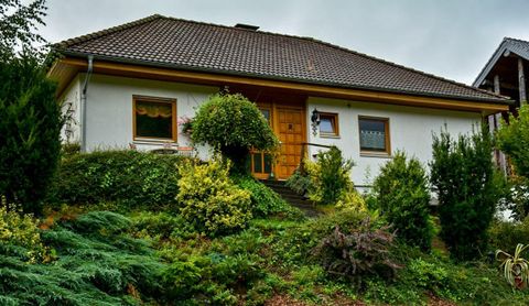 An inviting house on a hill, surrounded by green bushes and plants. The facade is light with a brown door and a cozy entrance area.