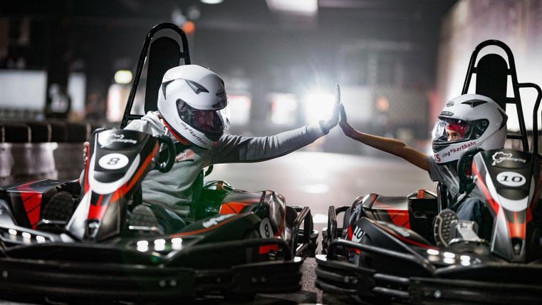 Zwei Kartfahrer in Rennanzügen geben sich ein High-Five. Die beiden Karts stehen nebeneinander in einer beleuchteten Rennhalle.