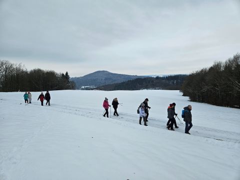 Een groep mensen wandelt in de sneeuw op een open veld. Op de achtergrond zijn bomen en zachte heuvels te zien.