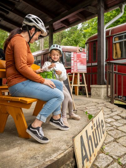 Two people wearing bicycle helmets are sitting at the Ahrhütte during the Tour de Ahrtal next to a red locomotive and a sign that reads “Passing Trains.”