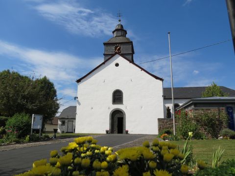Eine weiße Kirche mit einem charakteristischen Dach und einer Uhr. Blumenbeete umgeben das Gebäude und der Himmel ist klar blau.
